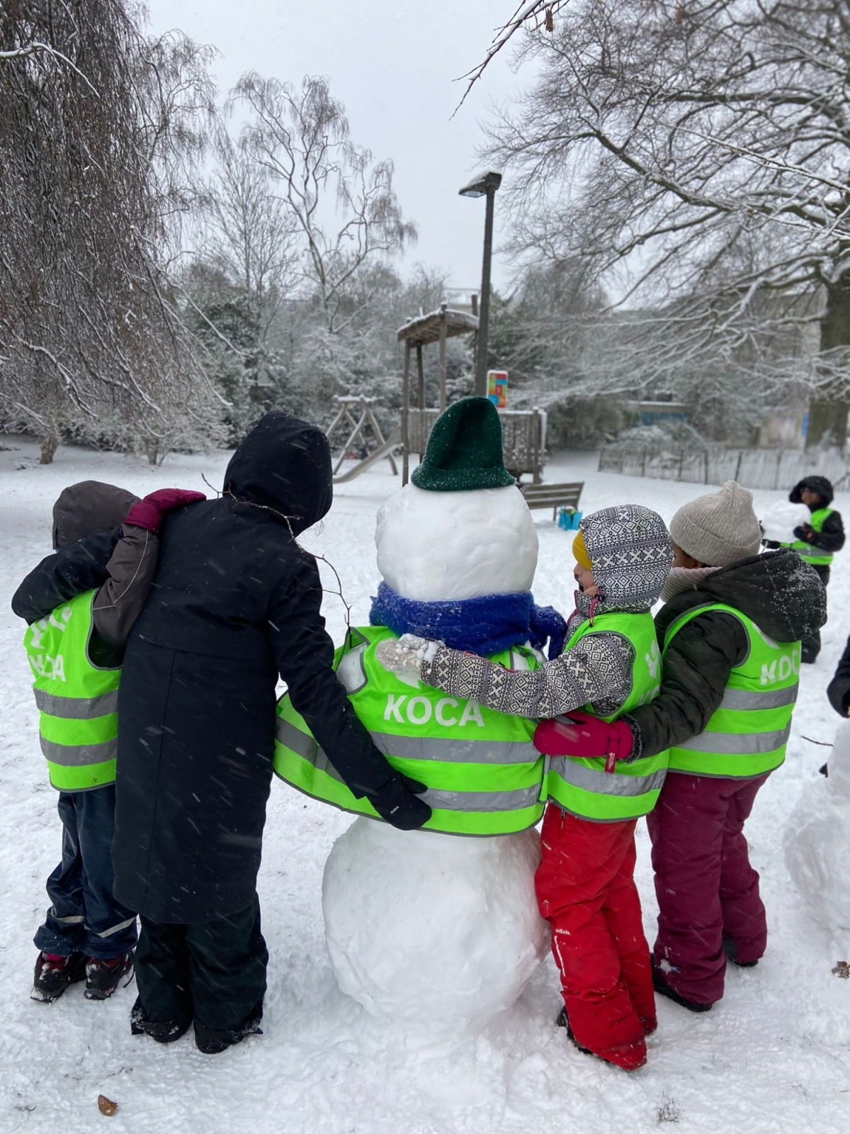 Drie kinderen en een leerkracht staan met de armen om elkaar. In het midden staat een sneeuwman met een fluovestje van de school.